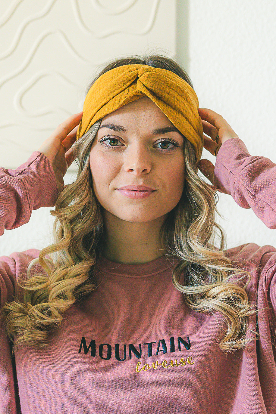 La photo montre une femme tenant un bandeau de couleur moutarde sur sa tête. Elle porte un pull rose avec l'inscription "MOUNTAIN" en lettres capitales de couleur foncée. Ses cheveux sont blonds et bouclés. Elle semble être devant un fond neutre avec un motif subtil. La photo est prise de la poitrine vers le haut.