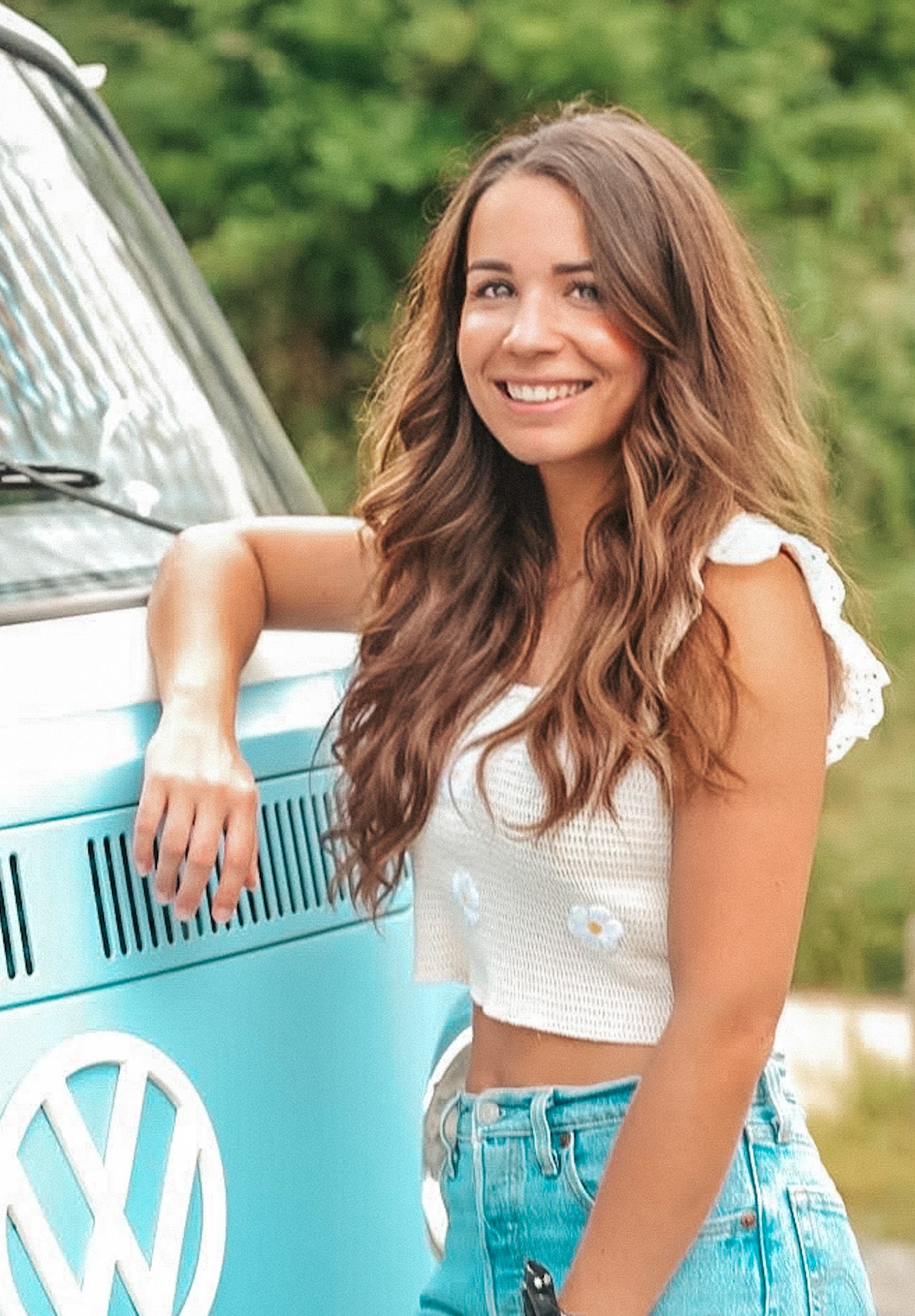 Sur la photo, il y a une jeune femme souriante avec des cheveux longs et ondulés de couleur châtain clair. Elle porte un haut court blanc à bretelles et un jean bleu délavé. Elle est appuyée contre un van bleu clair avec le logo de Volkswagen visible sur le devant. La photo est prise en extérieur avec de la végétation floue en arrière-plan.