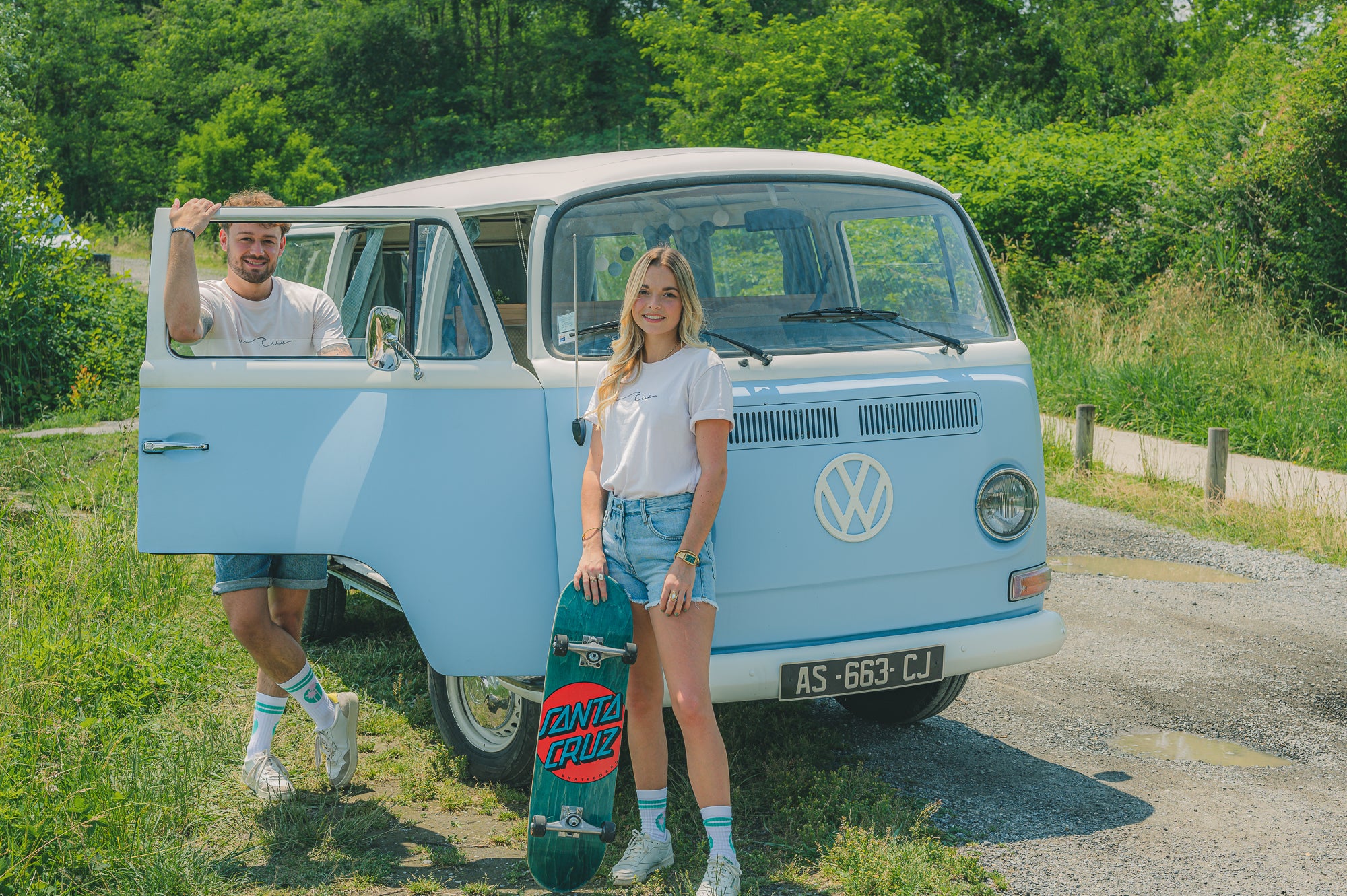 Sur la photo, il y a un homme et une femme debout à côté d'un van Volkswagen de couleur bleu pastel. L'homme est à gauche, il est appuyé sur la portière. La femme sur la droite tient un skateboard avec l'inscription "Santa Cruz". 