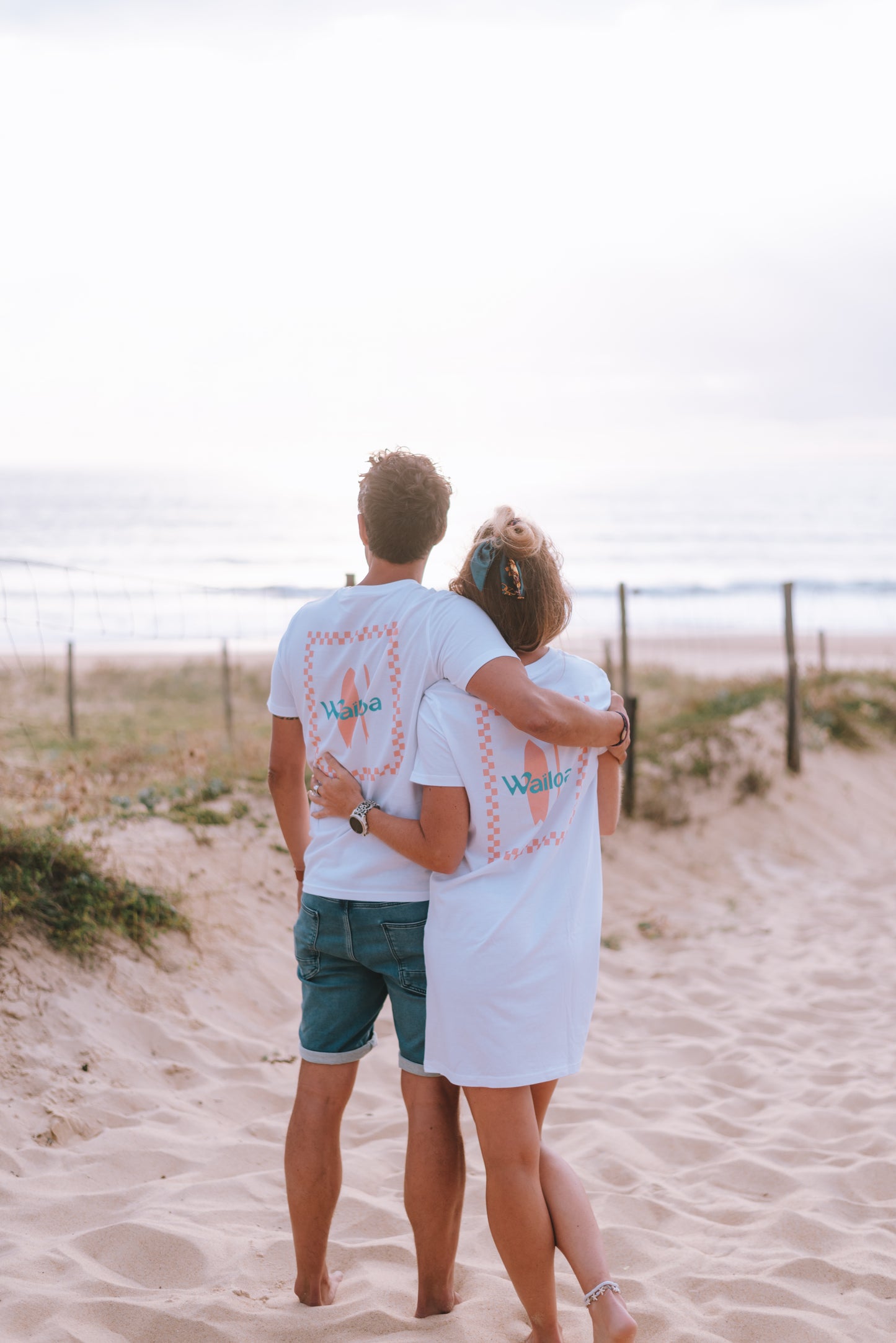 Couple sur la côte atlantique en bord de plage lors d'un coucher de soleil portant un t-shirt et une robe t-shirt assortis blanc et pêche. Un jolie dos à damier, un surf au centre et le logo de la marque Waïloa en bleu canard pour relever le tout.