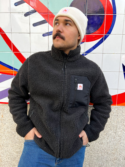 Il s'agit d'une photo d'un homme portant une veste polaire noire et un bonnet blanc. Il a une moustache et regarde vers le haut à sa droite. L'arrière-plan est un mur avec une peinture murale abstraite colorée, principalement en rouge, bleu et blanc.