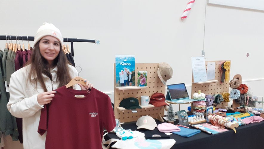La photo montre une femme souriante tenant un cintre avec un t-shirt bordeaux. Elle se trouve devant un stand avec divers articles exposés, comme des casquettes, des sacs et des bijoux.