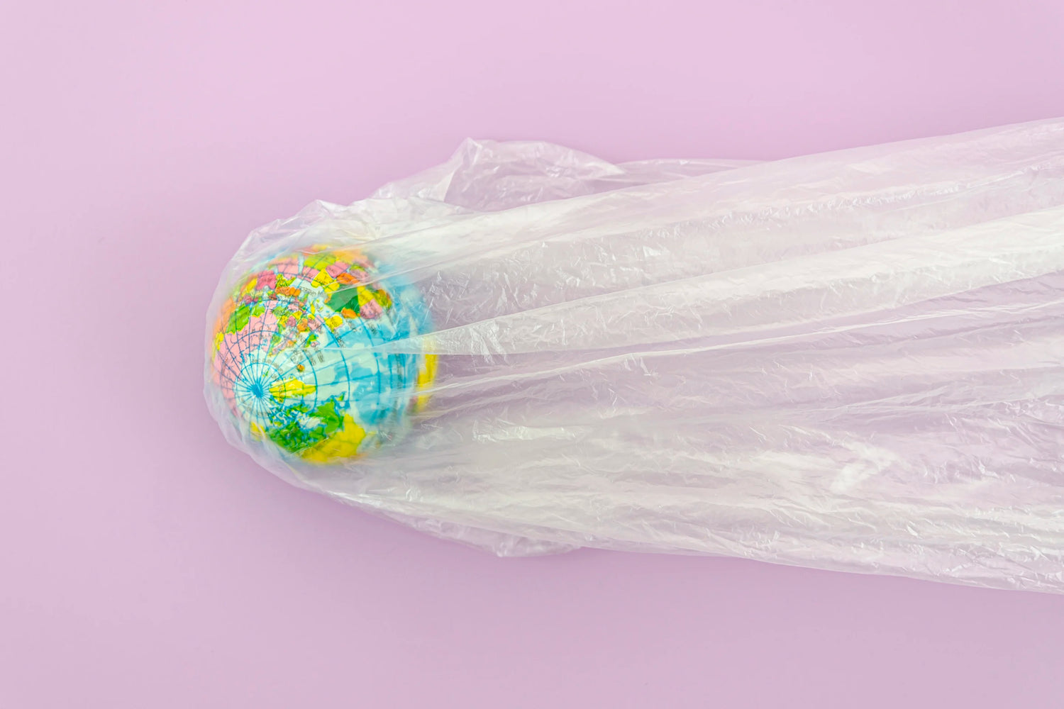 La photo montre un globe terrestre recouvert d'un sac en plastique transparent. Le globe est centré et le sac tombe en plis verticaux jusqu'au bas de l'image. Le fond est de couleur lilas.