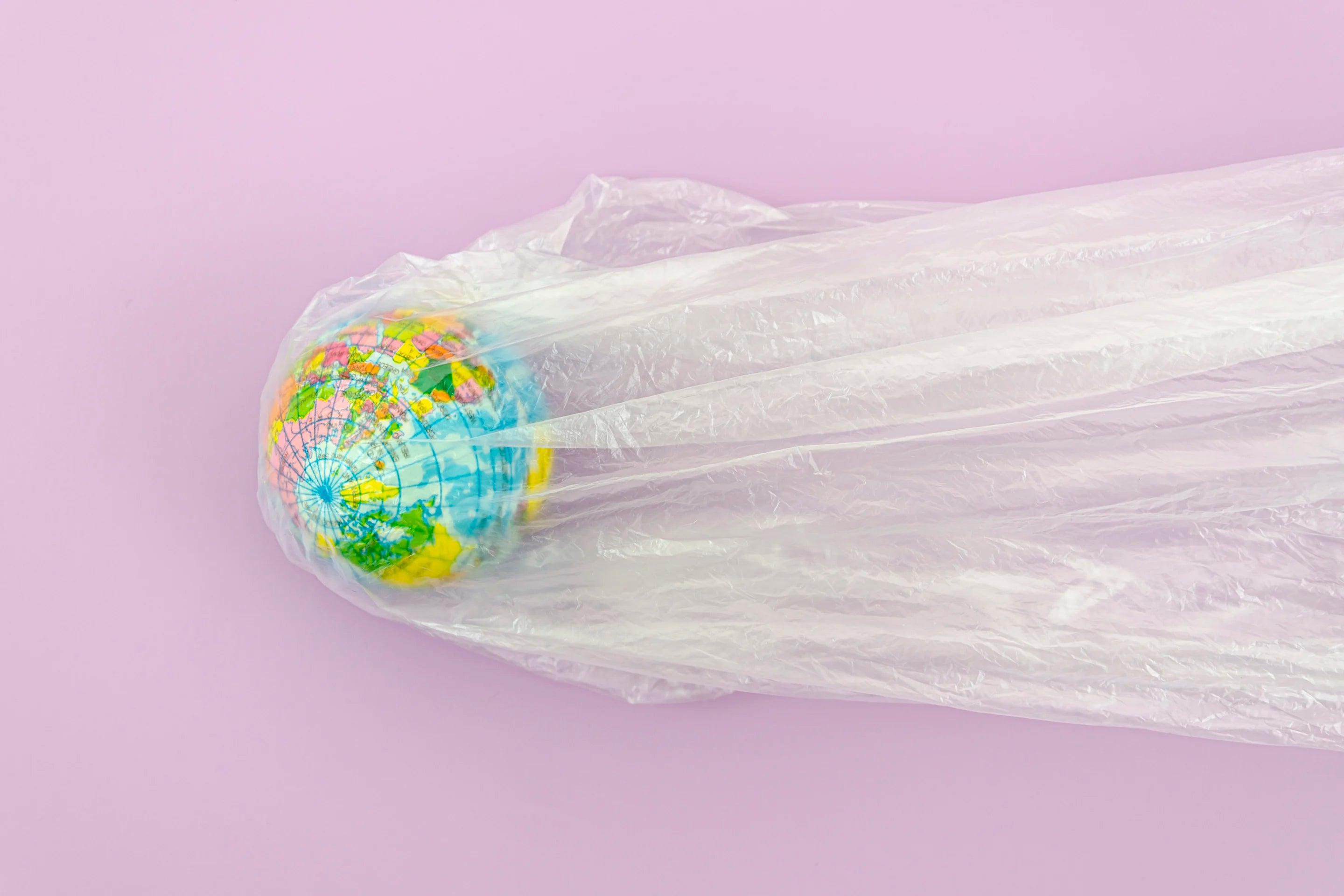 La photo montre un globe terrestre recouvert d'un sac en plastique transparent. Le globe est centré et le sac tombe en plis verticaux jusqu'au bas de l'image. Le fond est de couleur lilas.