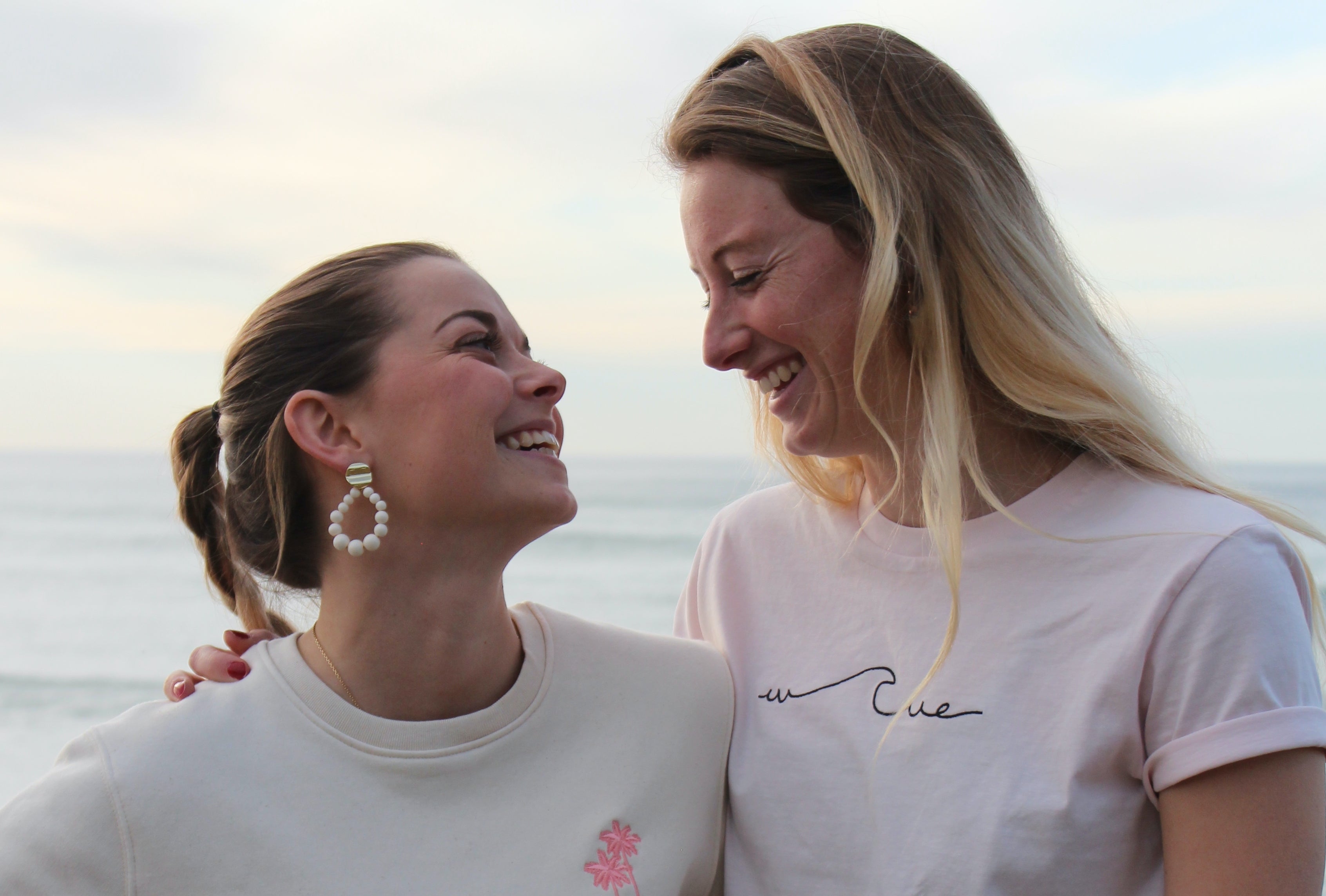 La photo montre deux femmes qui se font face et sourient largement. Elles semblent heureuses et complices. L'une porte des boucles d'oreilles en perles et un sweat écru avec unpalmier brodé côté coeur. Leurs cheveux sont blonds et lâchés pour celle de droite, coiffé en tresse pour celle de gauche. La photo est prise en bord de mer avec un fond flou clair.