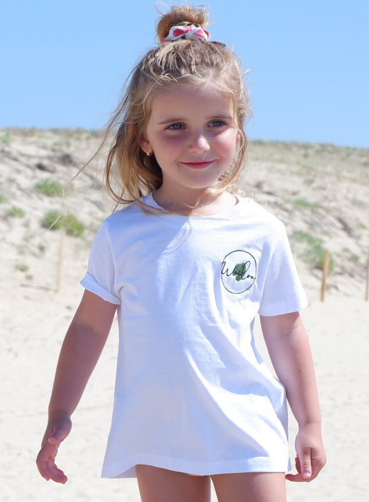 Il s'agit d'une jeune fille en tenue d'été, debout sur une plage non loin d'une dune de sable. Elle porte un t-shirt blanc avec un motif de monstera sur le côté gauche de la poitrine. Ses cheveux blonds sont partiellement attachés en haut avec un chouchou à fraises. Elle sourit et semble heureuse. L'arrière-plan est flou mais on peut distinguer voir une dune de sable.